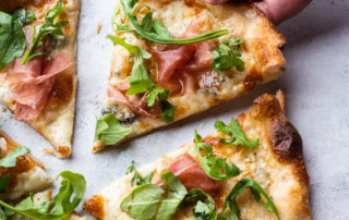 A hand reaches for for a slice of fresh pizza with prosciutto, arugula and balsamic vinegar