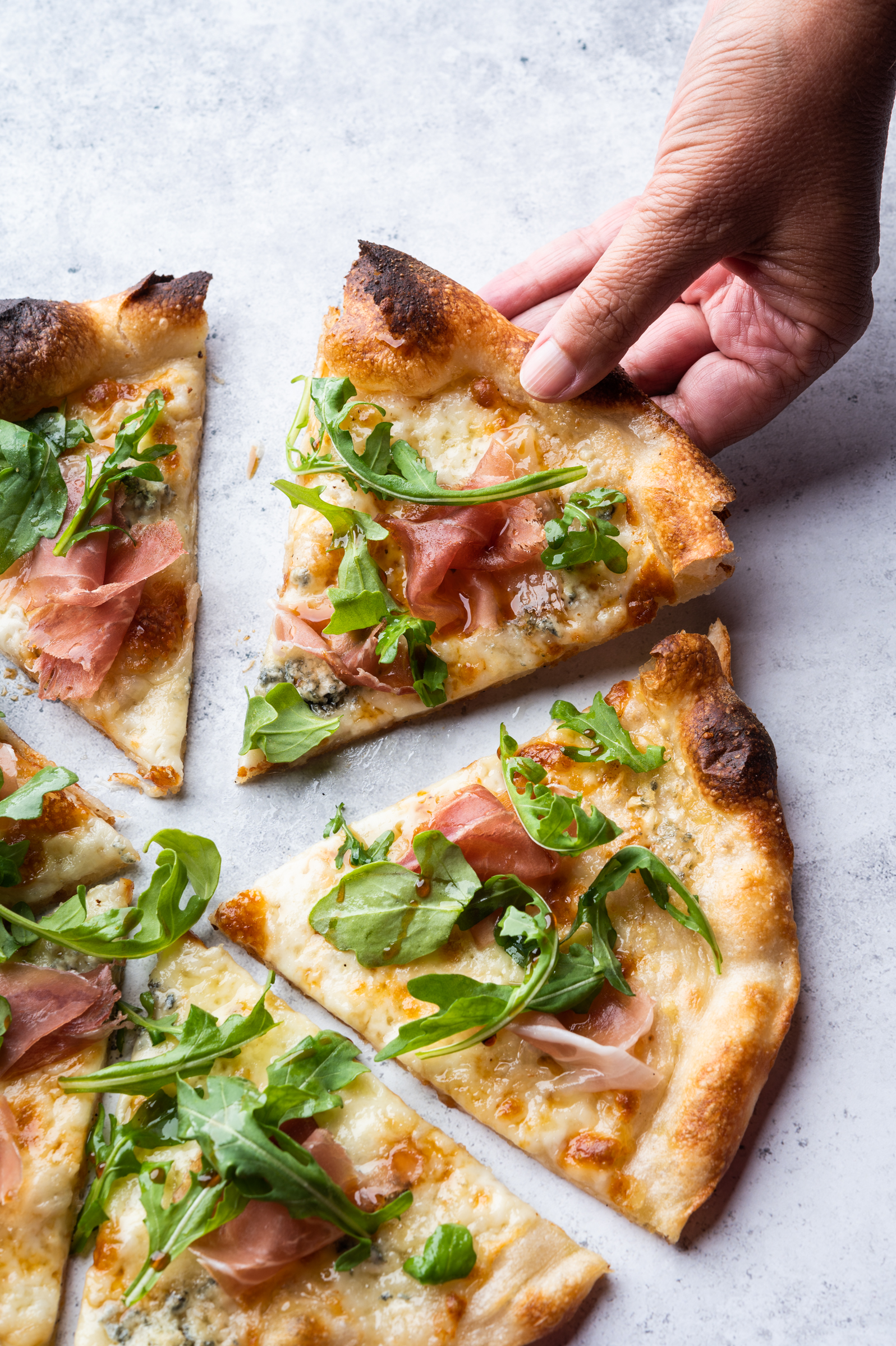A hand reaches for for a slice of fresh pizza with prosciutto, arugula and balsamic vinegar