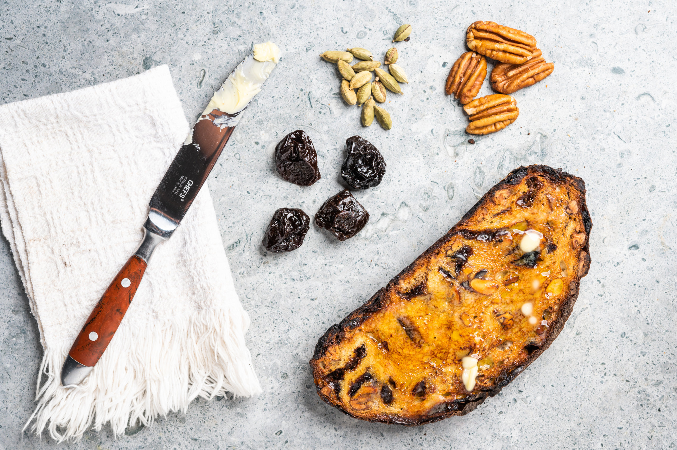 A butter knife next to toasted bread, California prunes, pecans and cardamom pods