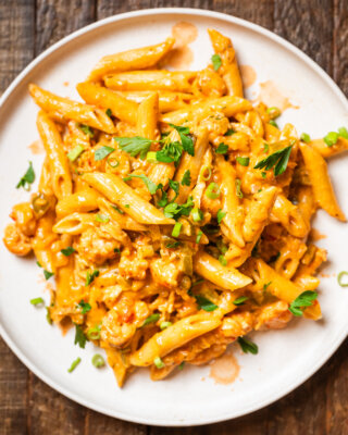 A plate of creamy crawfish penne pasta in a tomato-based sauce, garnished with chopped herbs and green onions.