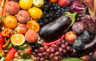 A variety of fruits and vegetables including grapes, peaches, peppers, pumpkins, cauliflower, and sweet potatoes are spread out in a colorful arrangement.