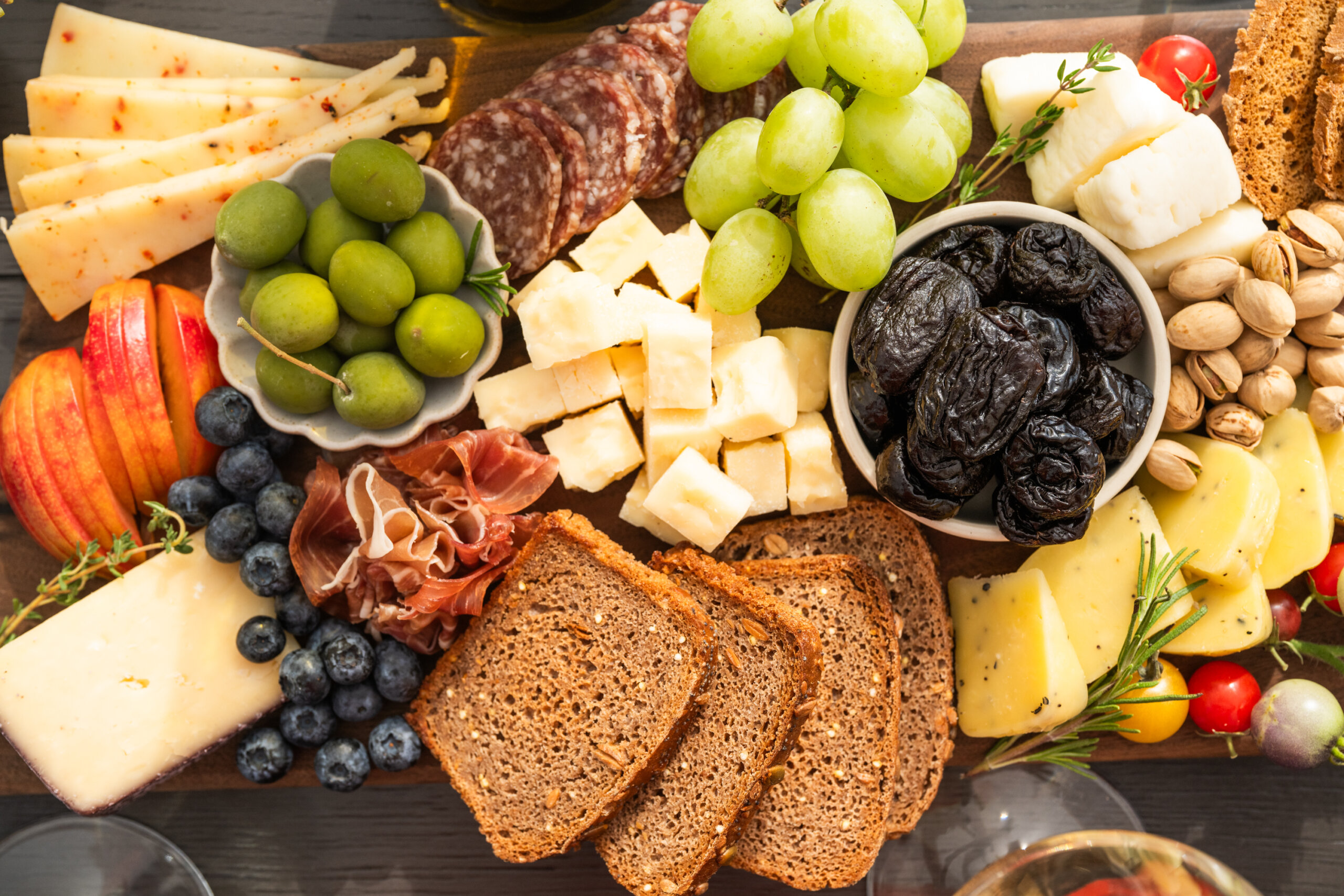 Top-down view of a cheeseboard featuring several cheeses, California prunes, pistachios, olives, grapes, and apricots.