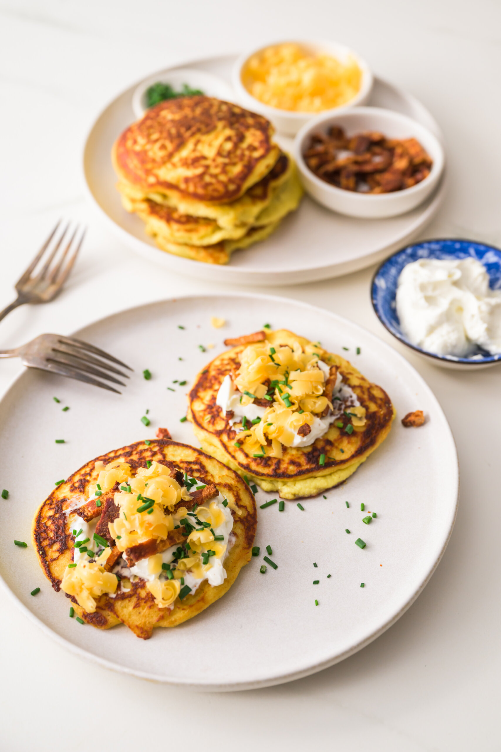 A plate with garnished savory ricotta pancakes in front of a plate filled with all the toppings.