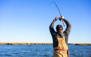 A fisherman holds his rod high to reel in a fish in the shallow marsh