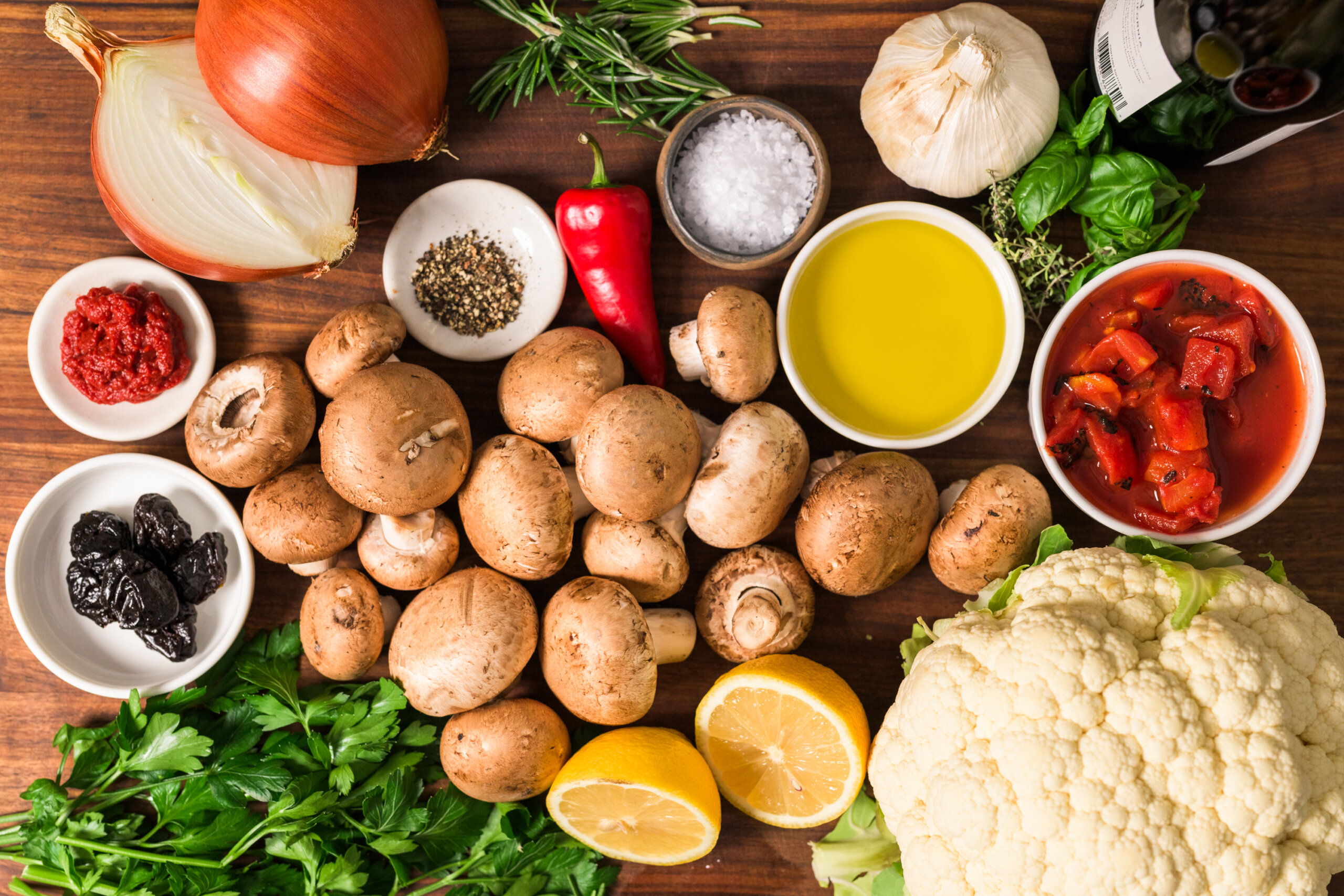 Assorted fresh ingredients on a wooden surface, including mushrooms, cauliflower, lemon, onion, herbs, spices, garlic, olive oil, tomatoes, red pepper, tomato paste, and olives.