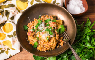 A bowl of vegetable bolognese topped with grated parmesan cheese and parsley. A fork rests on the edge. A cloth with a lemon pattern and a small bowl of salt are nearby on a wooden surface.