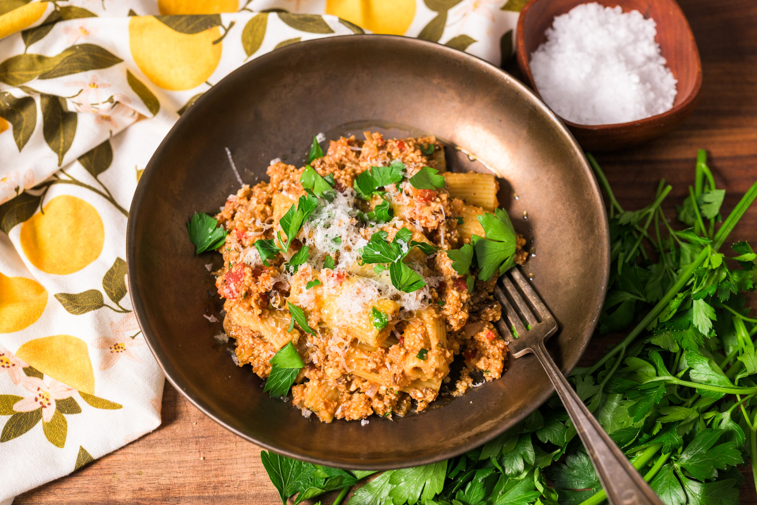 A bowl of vegetable bolognese topped with grated parmesan cheese and parsley. A fork rests on the edge. A cloth with a lemon pattern and a small bowl of salt are nearby on a wooden surface.