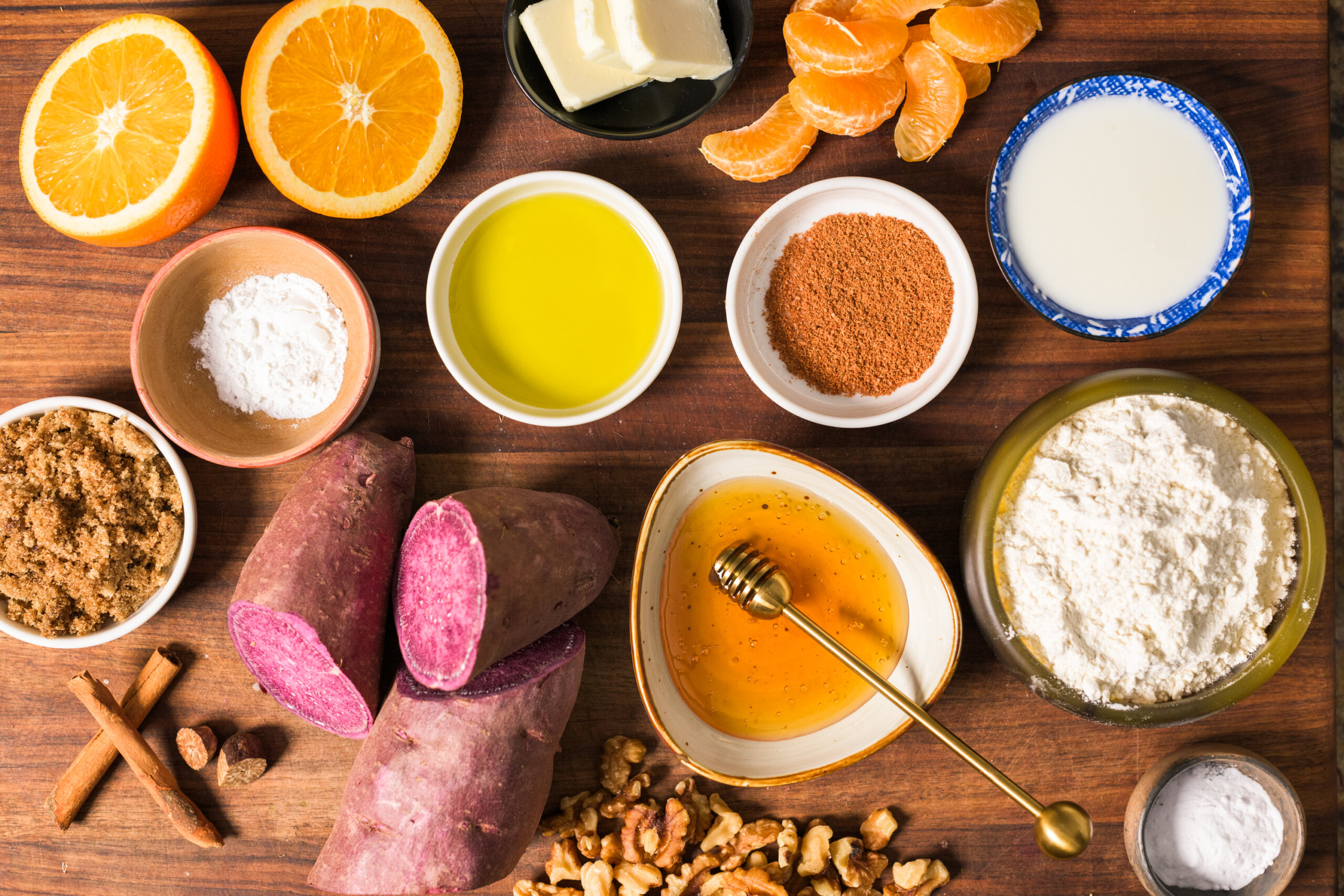 Various ingredients on a wooden table: sliced oranges, butter, milk, mandarin segments, flour, purple sweet potatoes, honey, oil, brown sugar, cinnamon sticks, nuts, cocoa powder.