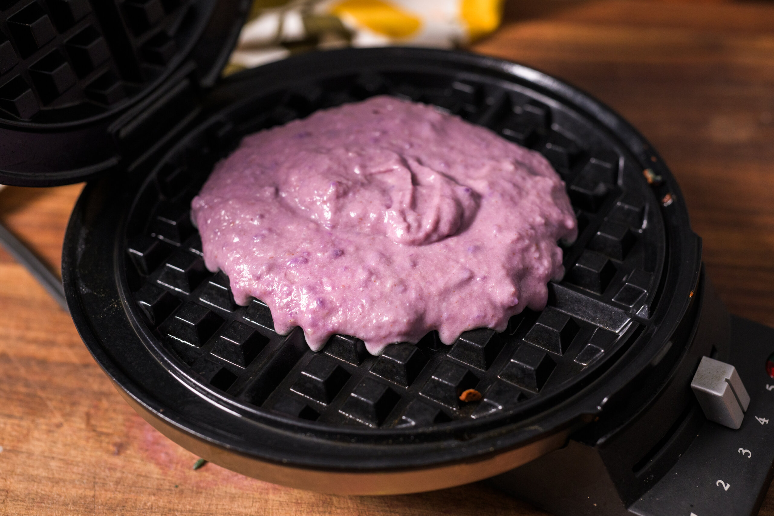 Purple waffle batter spread on a waffle iron, ready to cook, placed on a wooden surface.