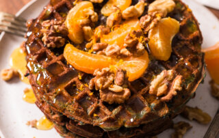 A stack of sweet potato waffles topped with mandarin orange slices, walnuts, and honey syrup on a white plate, with additional butter and a patterned cloth in the background.