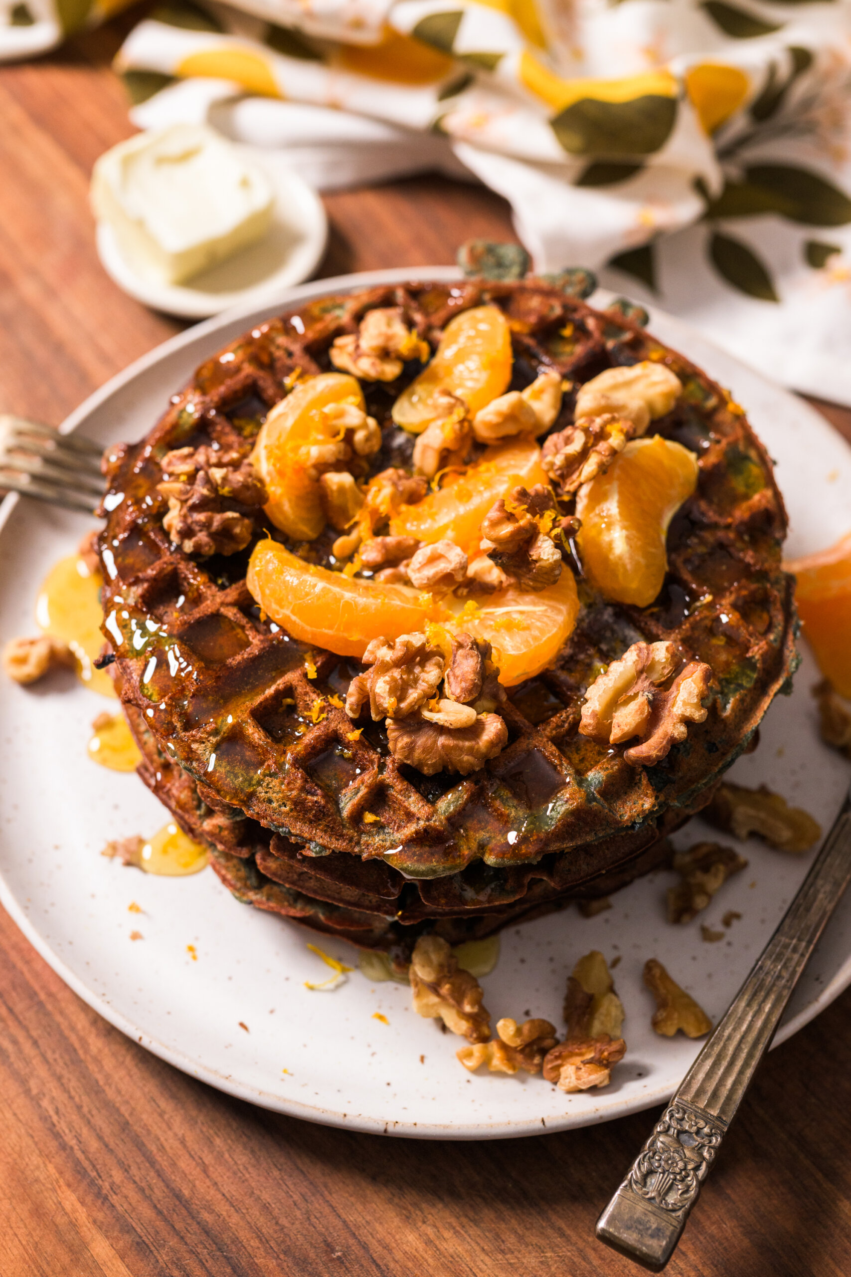 A stack of sweet potato waffles topped with mandarin orange slices, walnuts, and honey syrup on a white plate, with additional butter and a patterned cloth in the background.