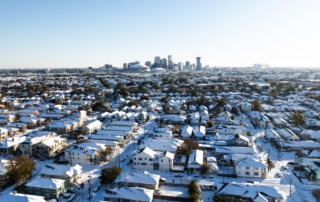 A snow-covered residential neighborhood with a city skyline in the background under a clear blue sky.