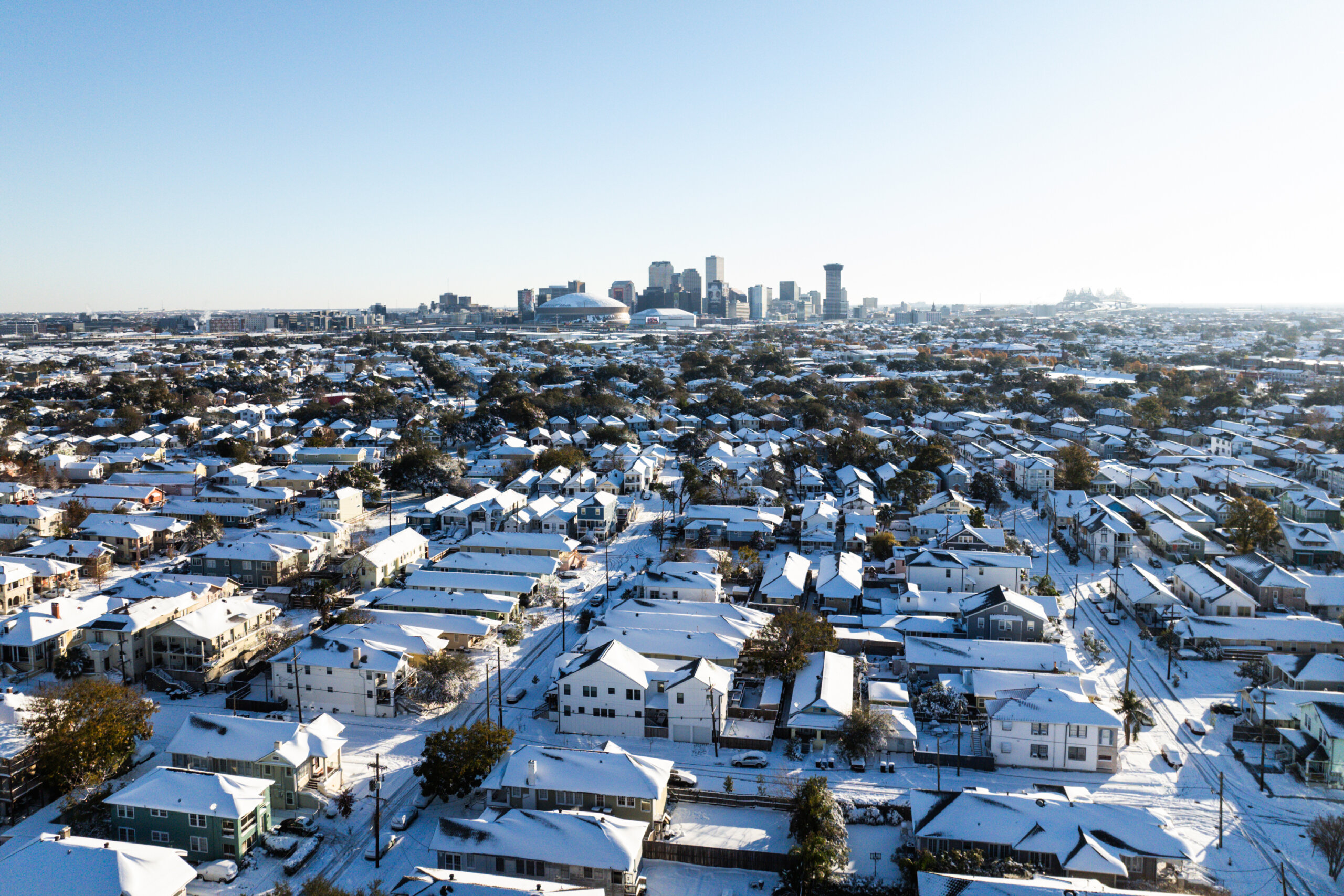 A snow-covered residential neighborhood with a city skyline in the background under a clear blue sky.