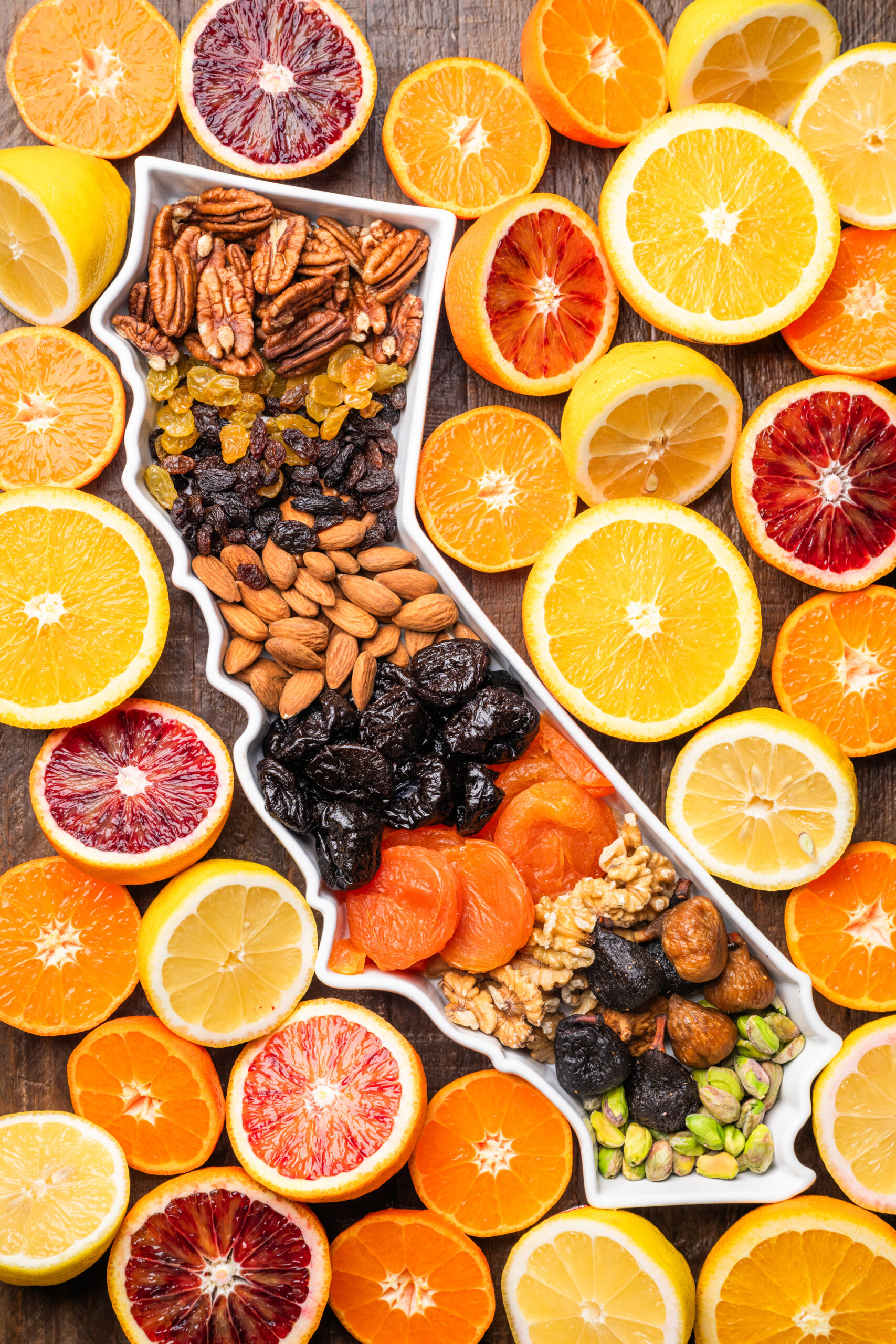 A platter with assorted nuts and dried fruits is surrounded by sliced citrus fruits, including oranges, lemons, and grapefruits, arranged on a wooden surface.