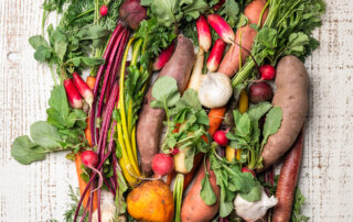 An assortment of fresh vegetables, including carrots, radishes, sweet potatoes, garlic, and leafy greens, arranged on a white wooden surface.