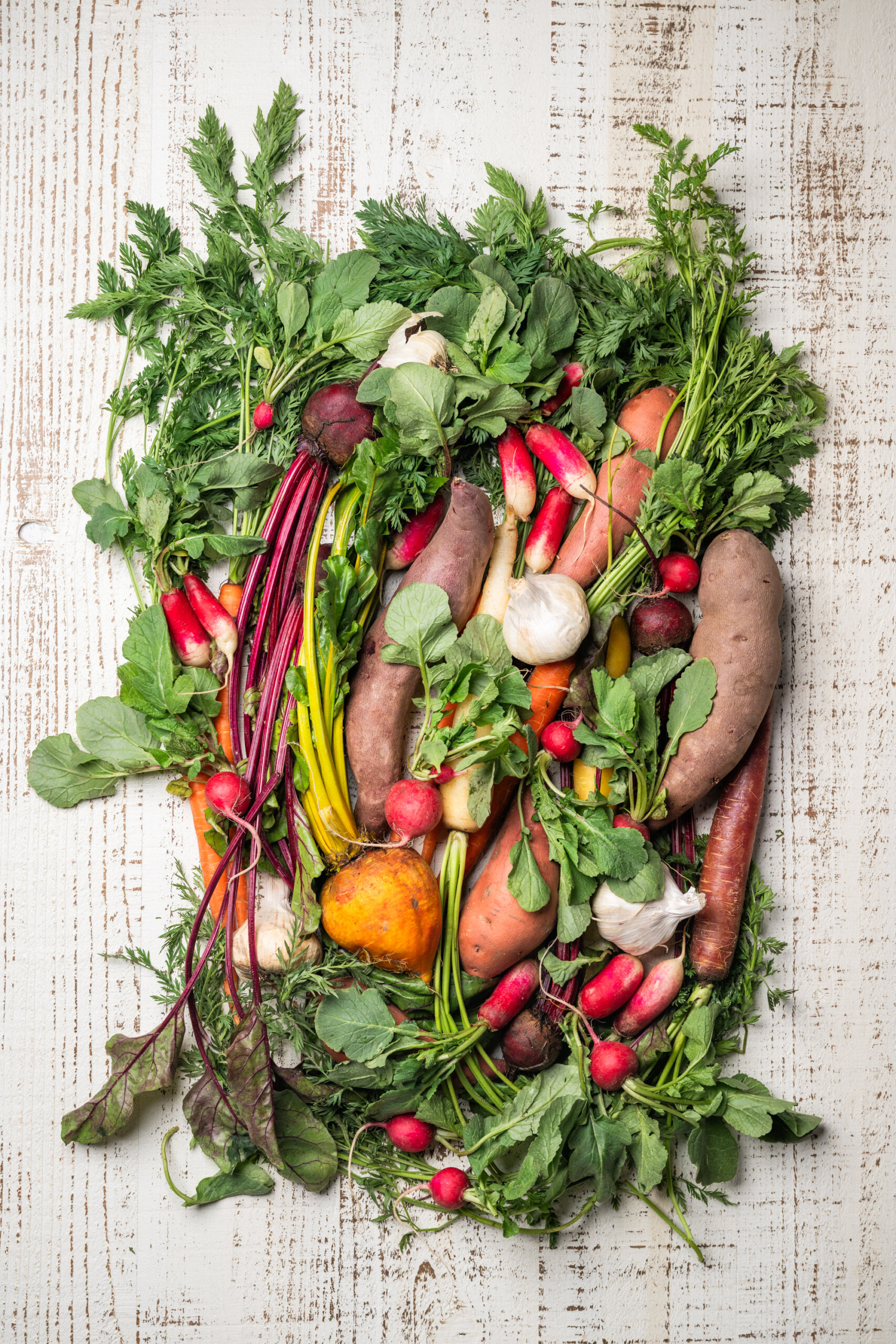 An assortment of fresh vegetables, including carrots, radishes, sweet potatoes, garlic, and leafy greens, arranged on a white wooden surface.