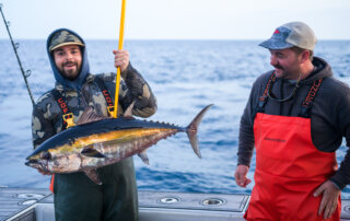 Two men on a boat, one holding a large fish with a fishing rod. Both are dressed in fishing gear, with the ocean in the background.