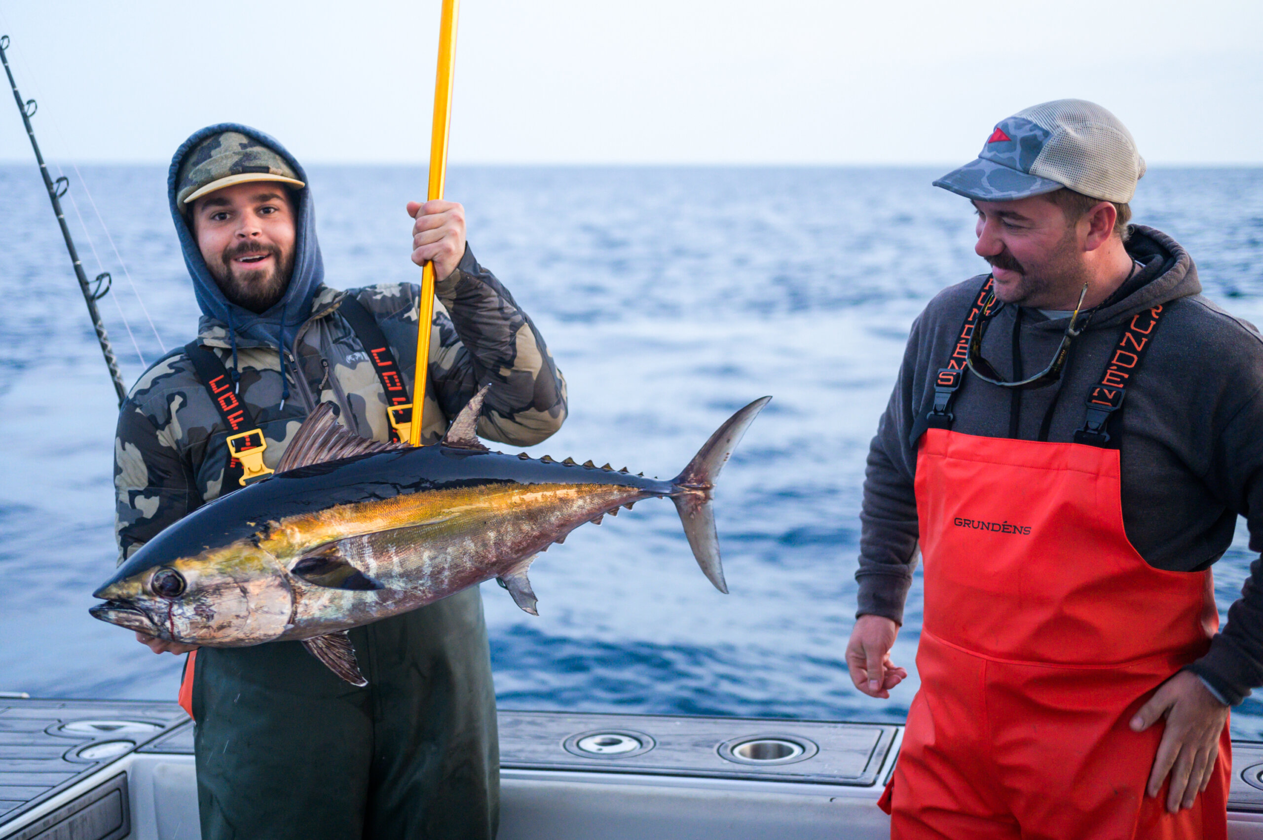 Two men on a boat, one holding a large fish with a fishing rod. Both are dressed in fishing gear, with the ocean in the background.