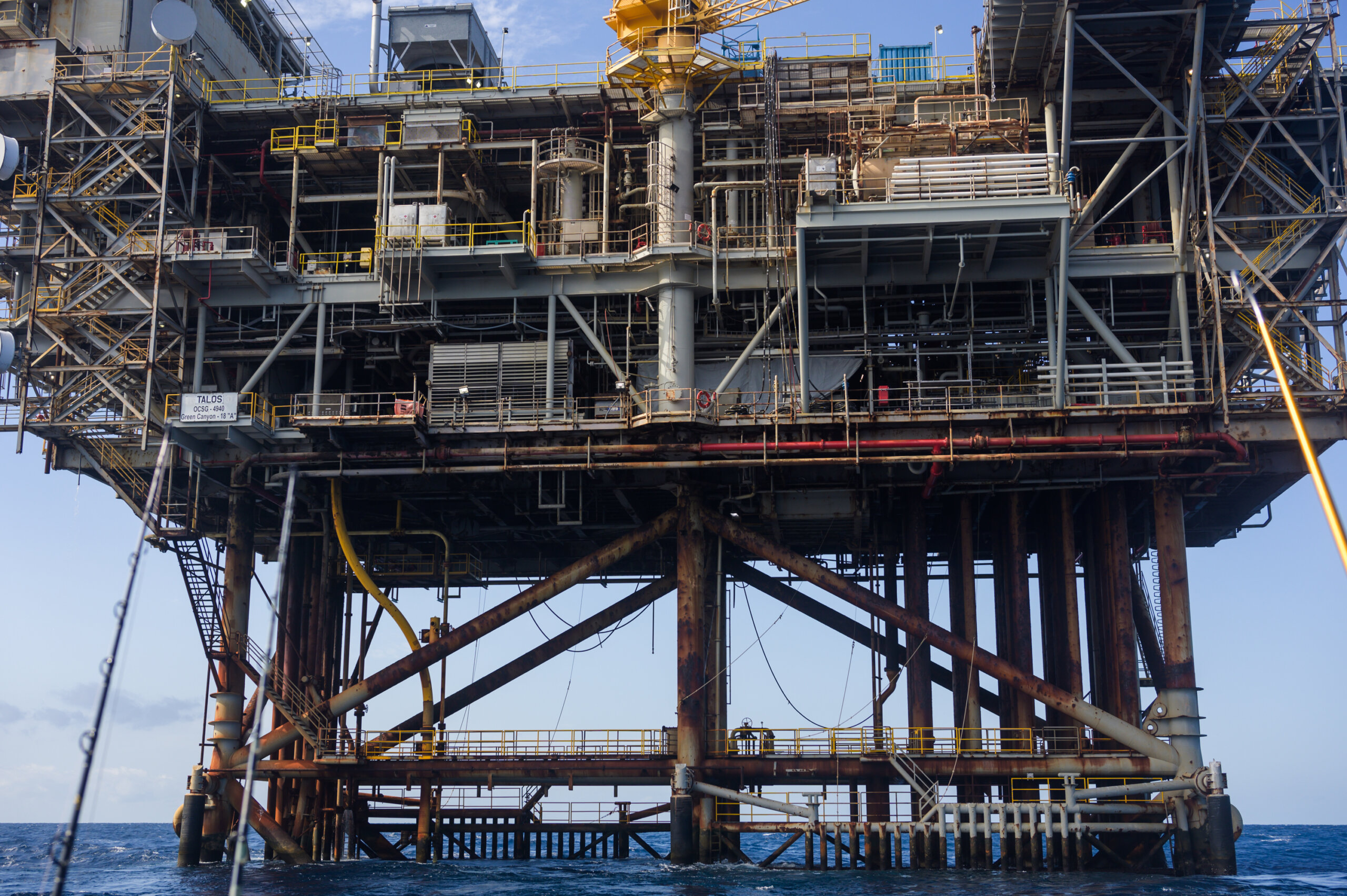 Offshore oil platform with complex steel structures rises above the ocean, supported by multiple legs. Sky visible in the background.