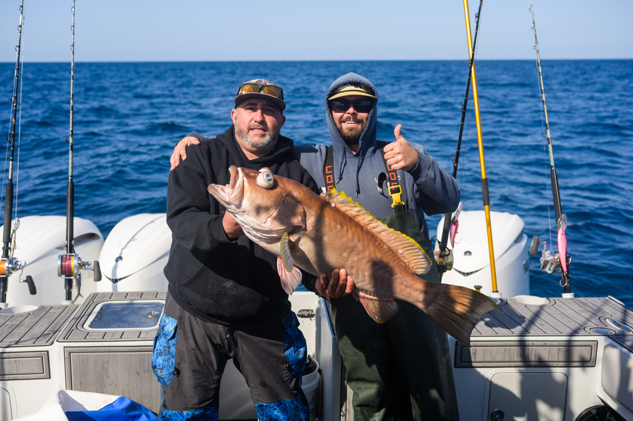 Two men on a boat holding a large fish, one man giving a thumbs-up. Fishing rods are visible in the background against a blue ocean.