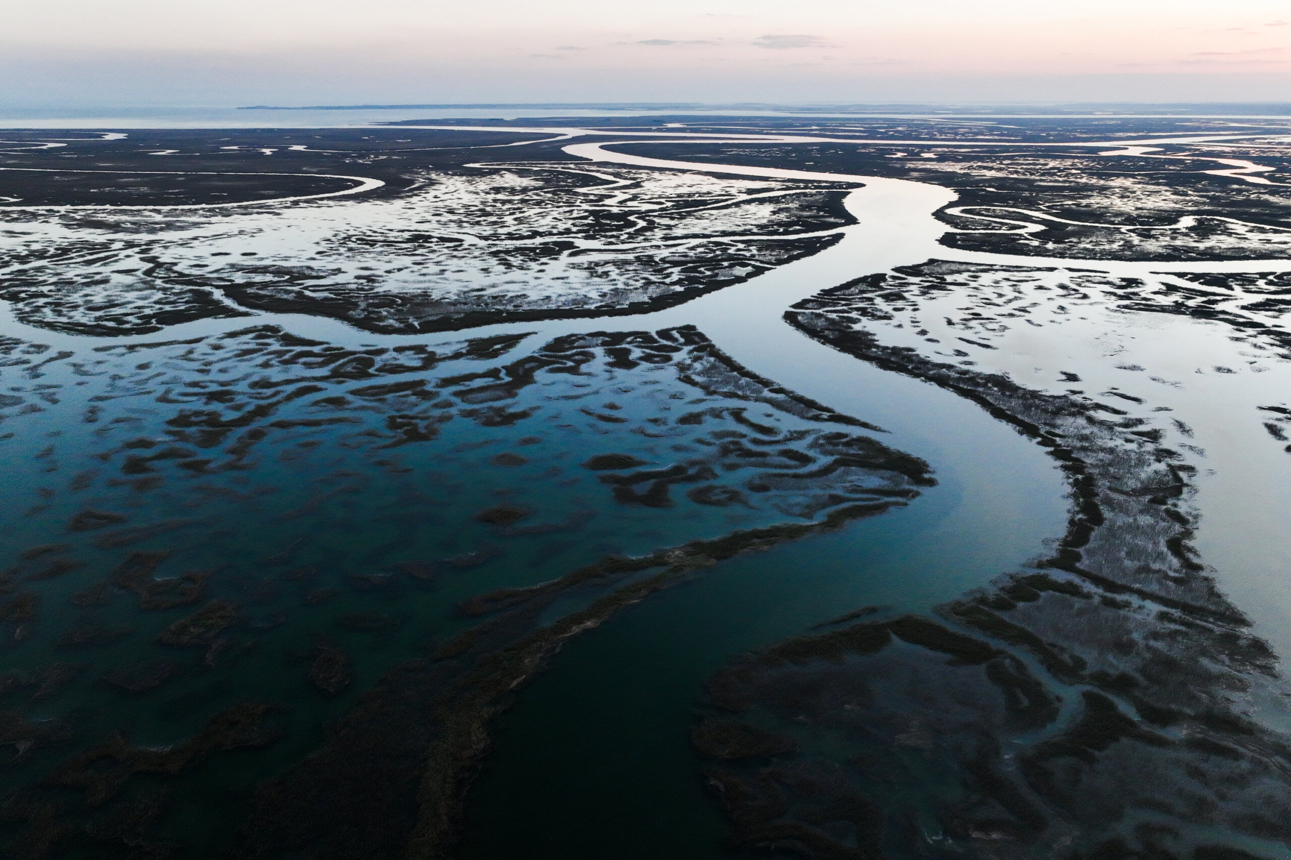 Aerial view of winding river channels flowing through a vast, marshy landscape under a blue sky at dusk.