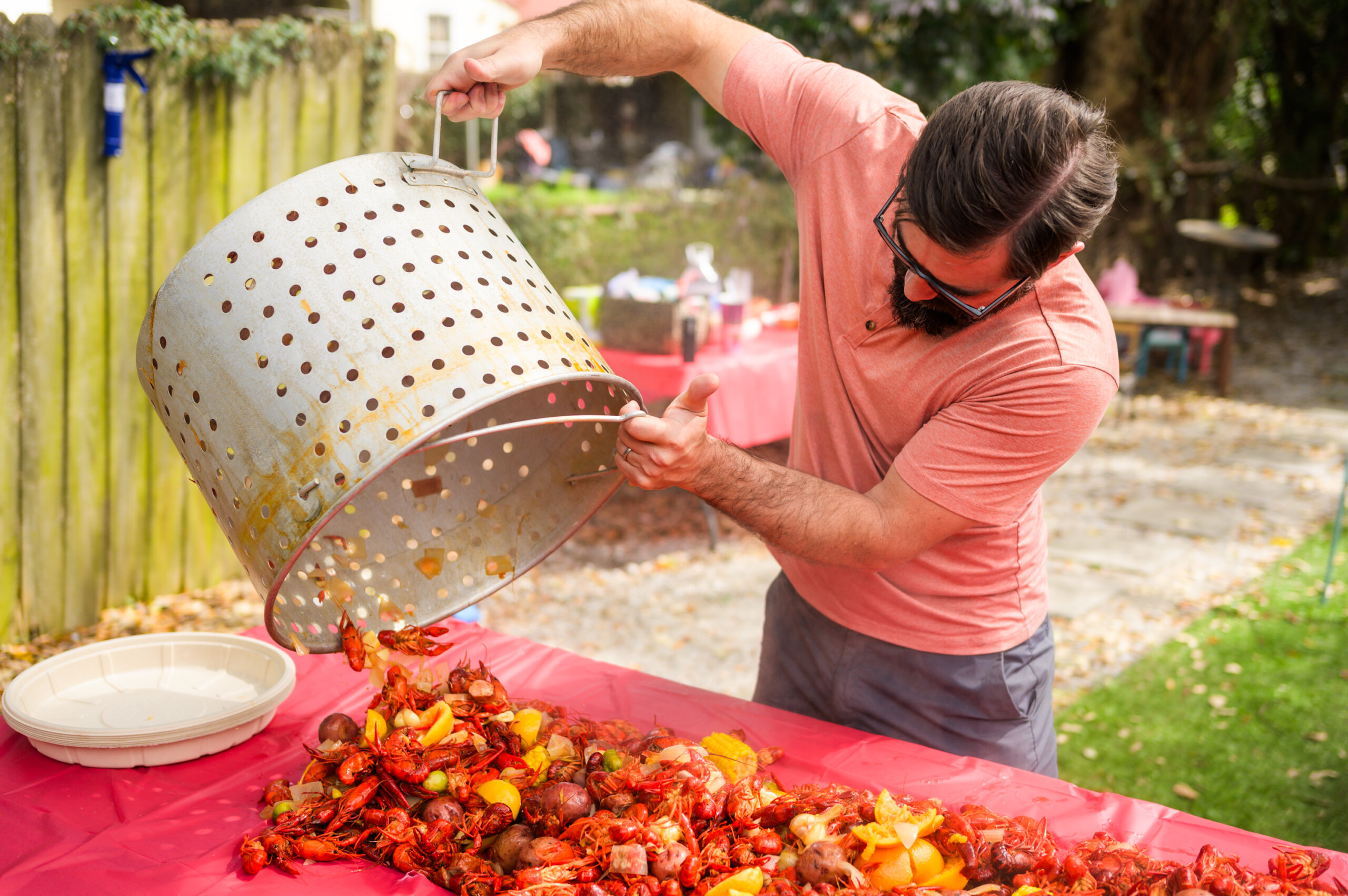 A man pours cooked crawfish, corn, and potatoes from a large boil pot onto a red table outdoors.