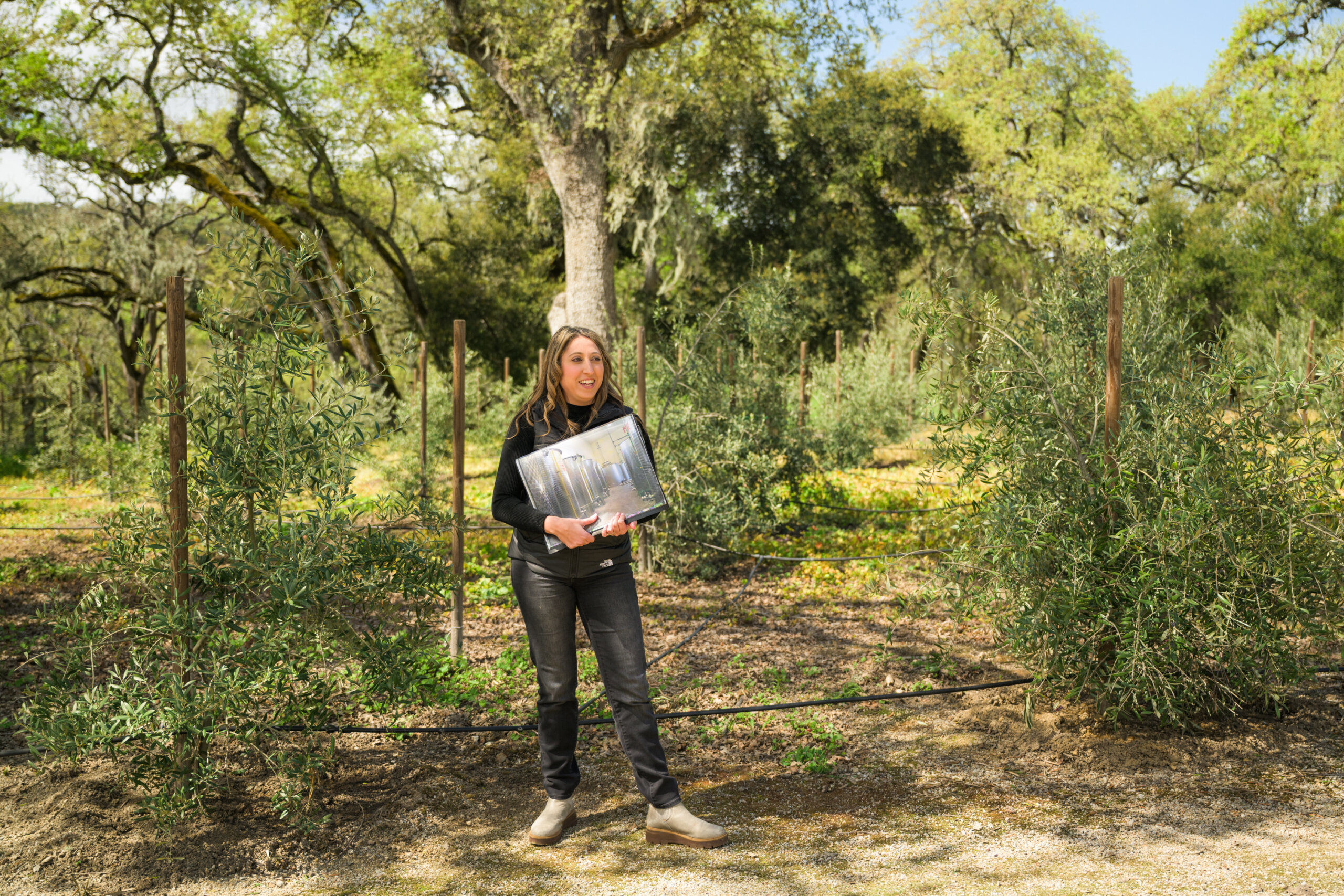A woman stands outdoors in an olive grove, holding a photo album. Trees and greenery are visible in the background under a sunny sky.