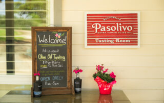 A chalkboard sign advertises olive oil tasting hours next to potted flowers and a red Pasolivo Tasting Room sign on a cream wall.