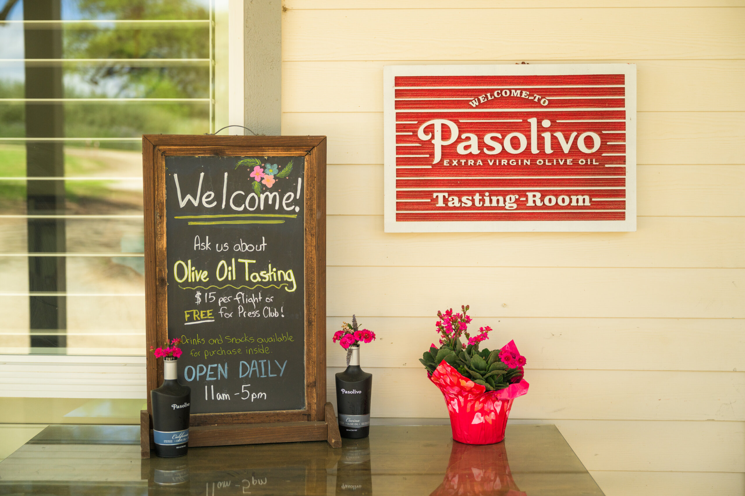 A chalkboard sign advertises olive oil tasting hours next to potted flowers and a red Pasolivo Tasting Room sign on a cream wall.