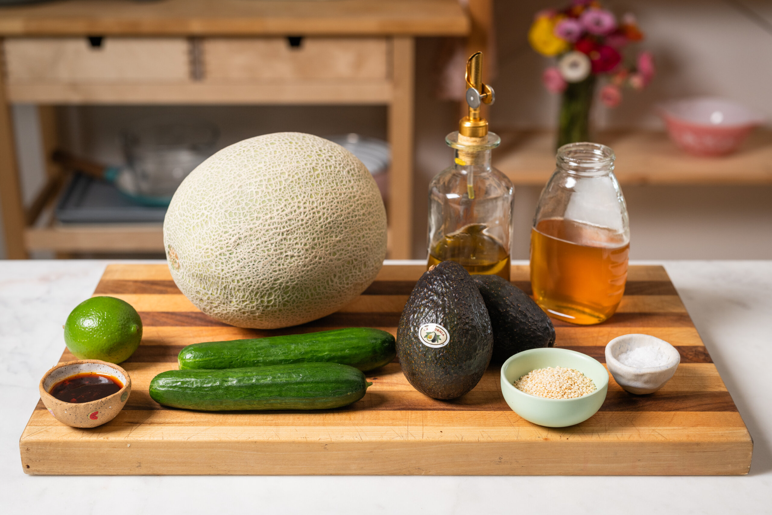 A wooden cutting board with a cantaloupe, two avocados, two cucumbers, a lime, sesame seeds, salt, oil, vinegar, and a small bowl of sauce. A kitchen in the background.