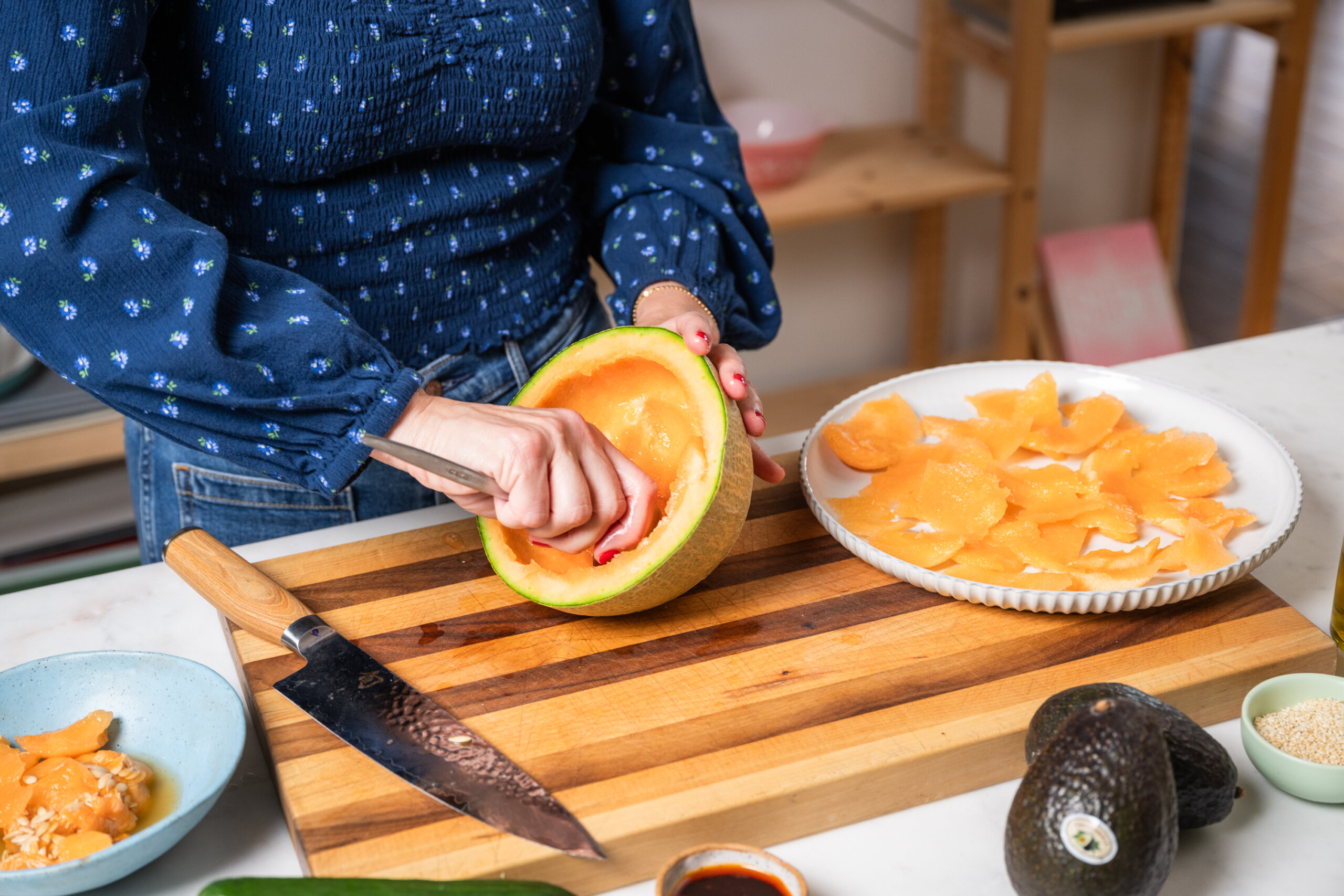 A person scoops out the flesh of a cantaloupe with a spoon on a wooden cutting board, with sliced cantaloupe, a knife, and other ingredients nearby.