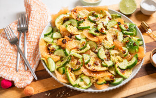 A plate of sliced cucumber, avocado and cantaloupe, sprinkled with sesame seeds, with lime halves and dressing ingredients in the background.