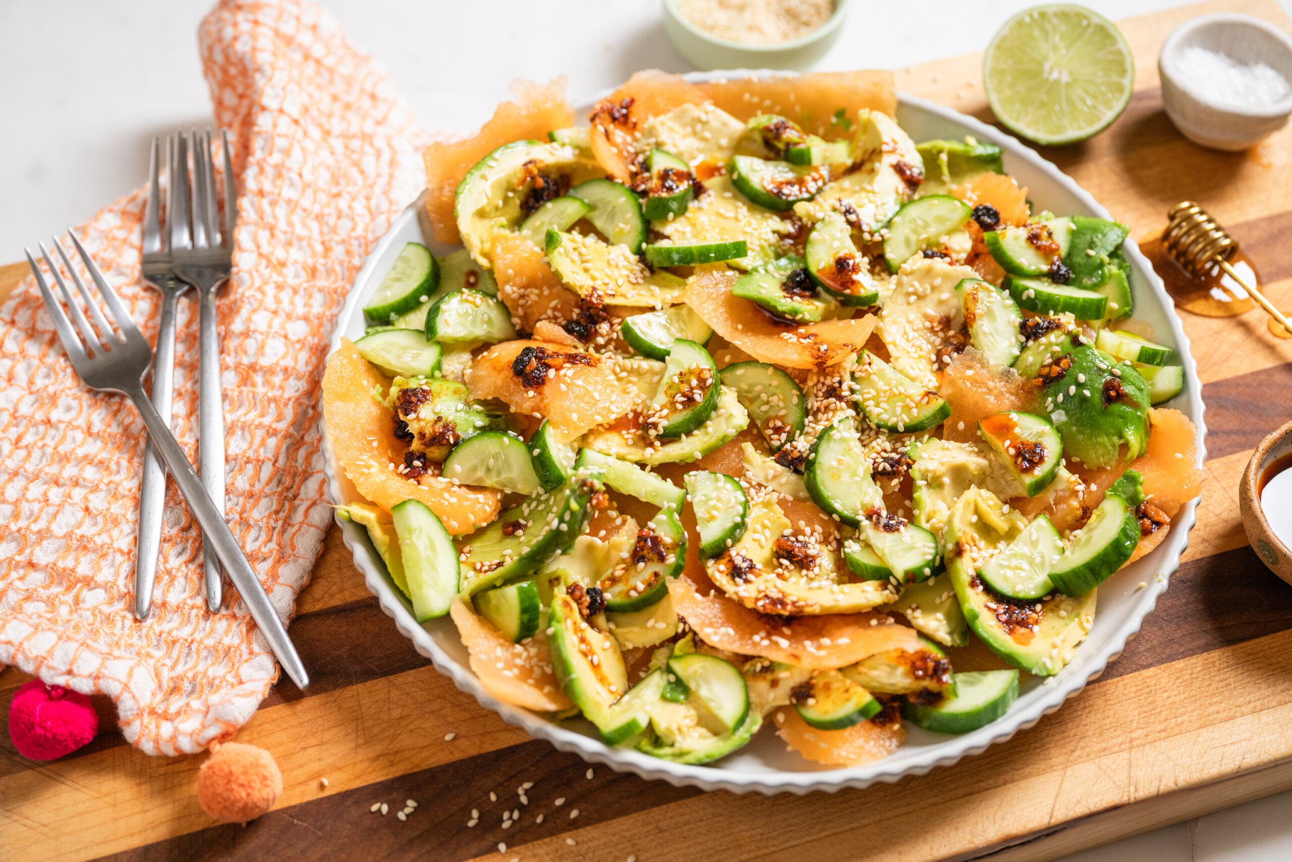 A plate of sliced cucumber, avocado and cantaloupe, sprinkled with sesame seeds, with lime halves and dressing ingredients in the background.