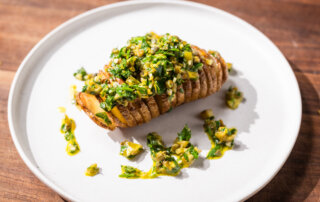 A sliced baked potato topped with a green and yellow herb sauce is served on a white plate on a wooden surface.
