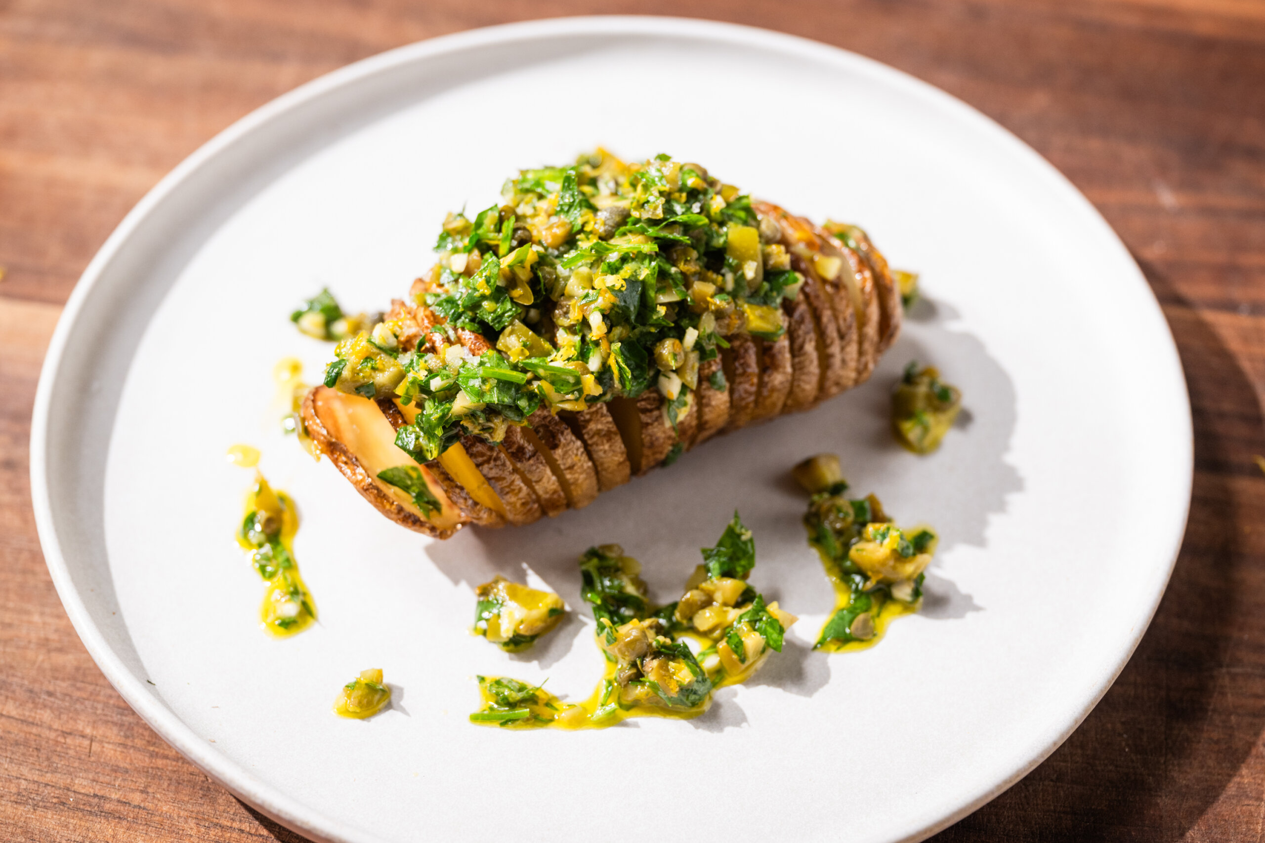 A sliced baked potato topped with a green and yellow herb sauce is served on a white plate on a wooden surface.