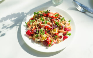 A white plate with a salad of strawberries, microgreens, fennel slices, marcona almonds, and tomatoes on a white table with sunlight and silverware nearby.