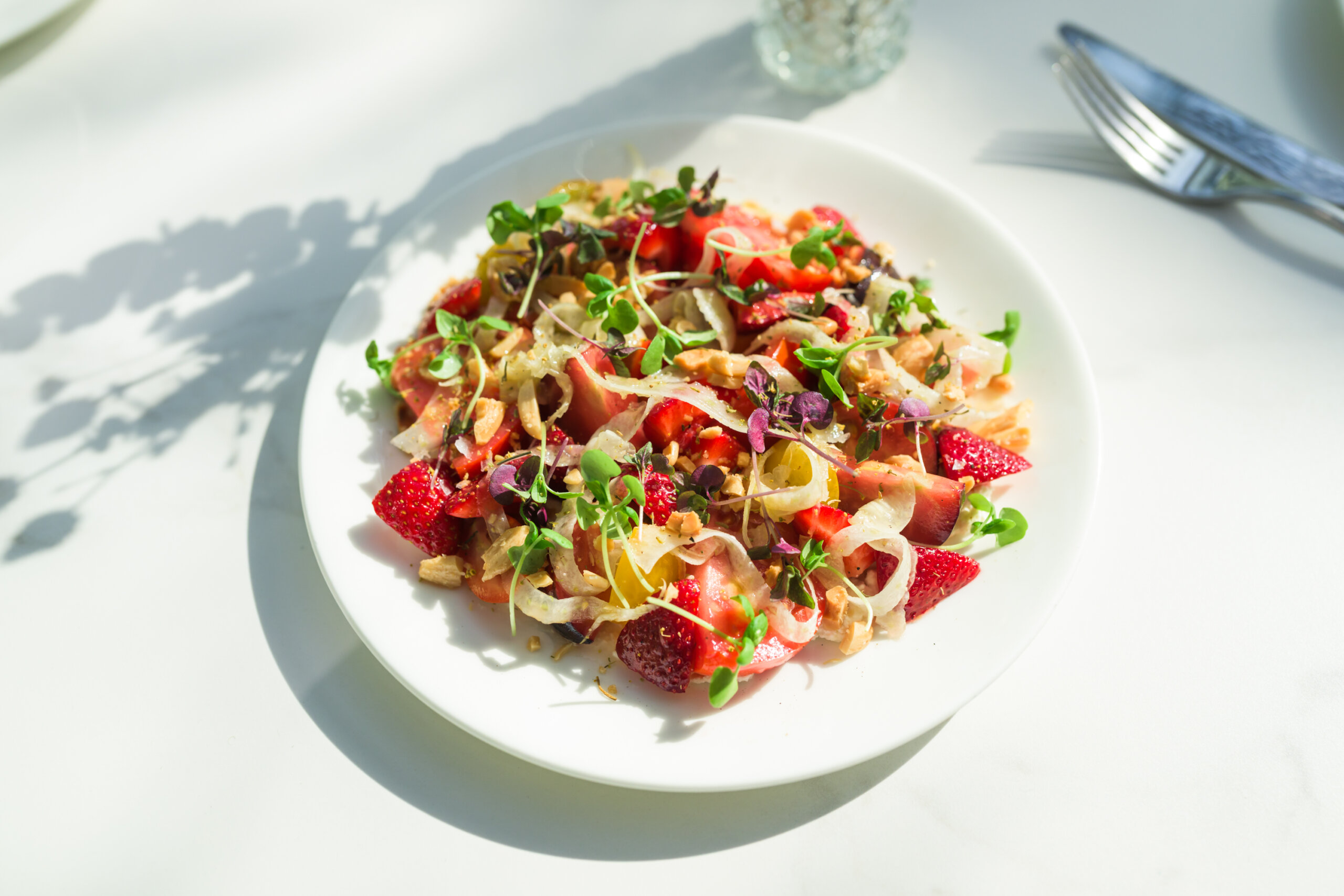 A white plate with a salad of strawberries, microgreens, fennel slices, marcona almonds, and tomatoes on a white table with sunlight and silverware nearby.