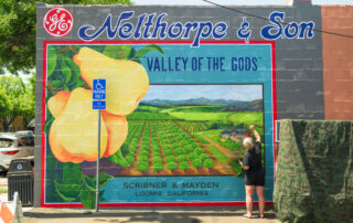 A person paints a mural on a wall that displays fruit, farmland, and the text “Valley of the Gods,” with a sign indicating handicapped parking nearby.
