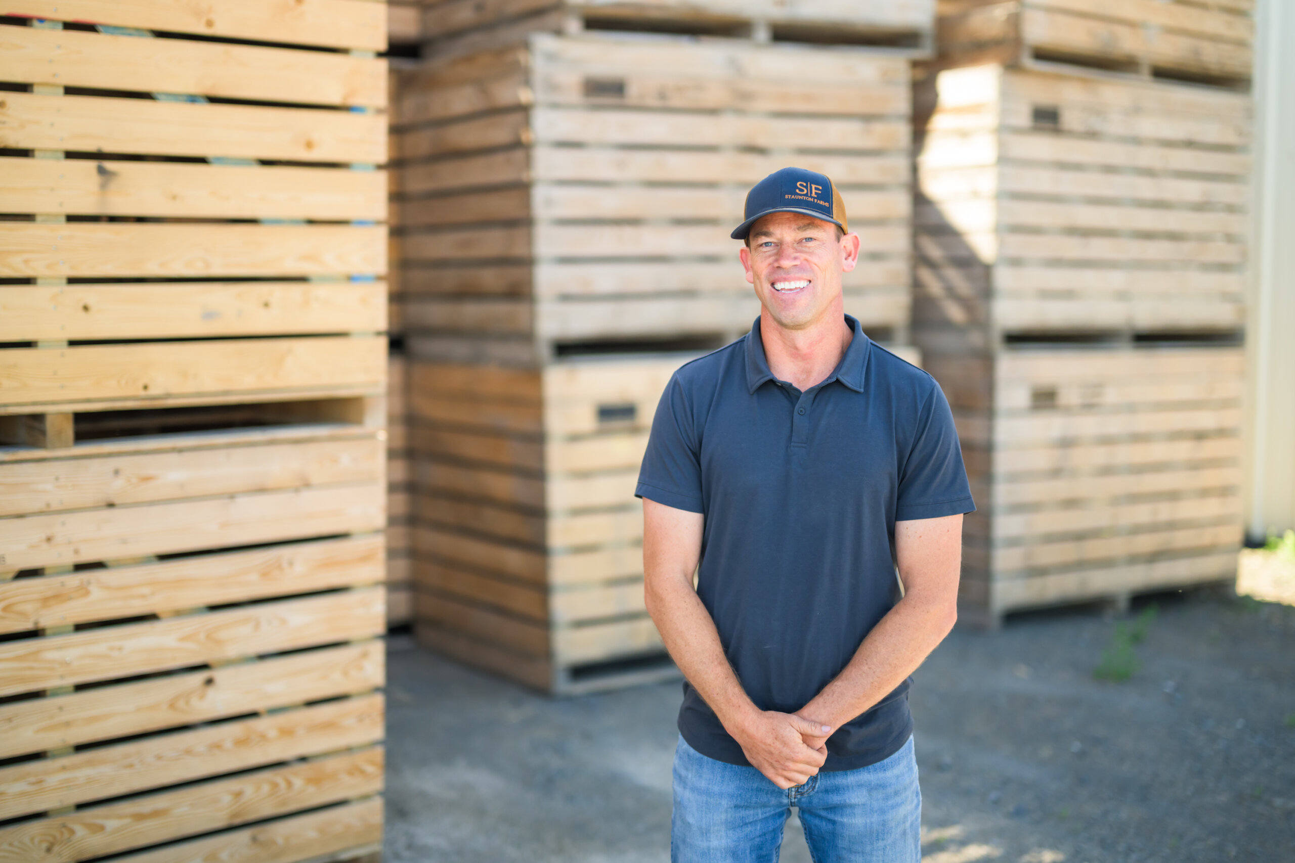 A man wearing a black polo shirt and cap stands in front of stacked wooden crates outdoors. A man wearing a black polo shirt and cap stands in front of stacked wooden crates outdoors.