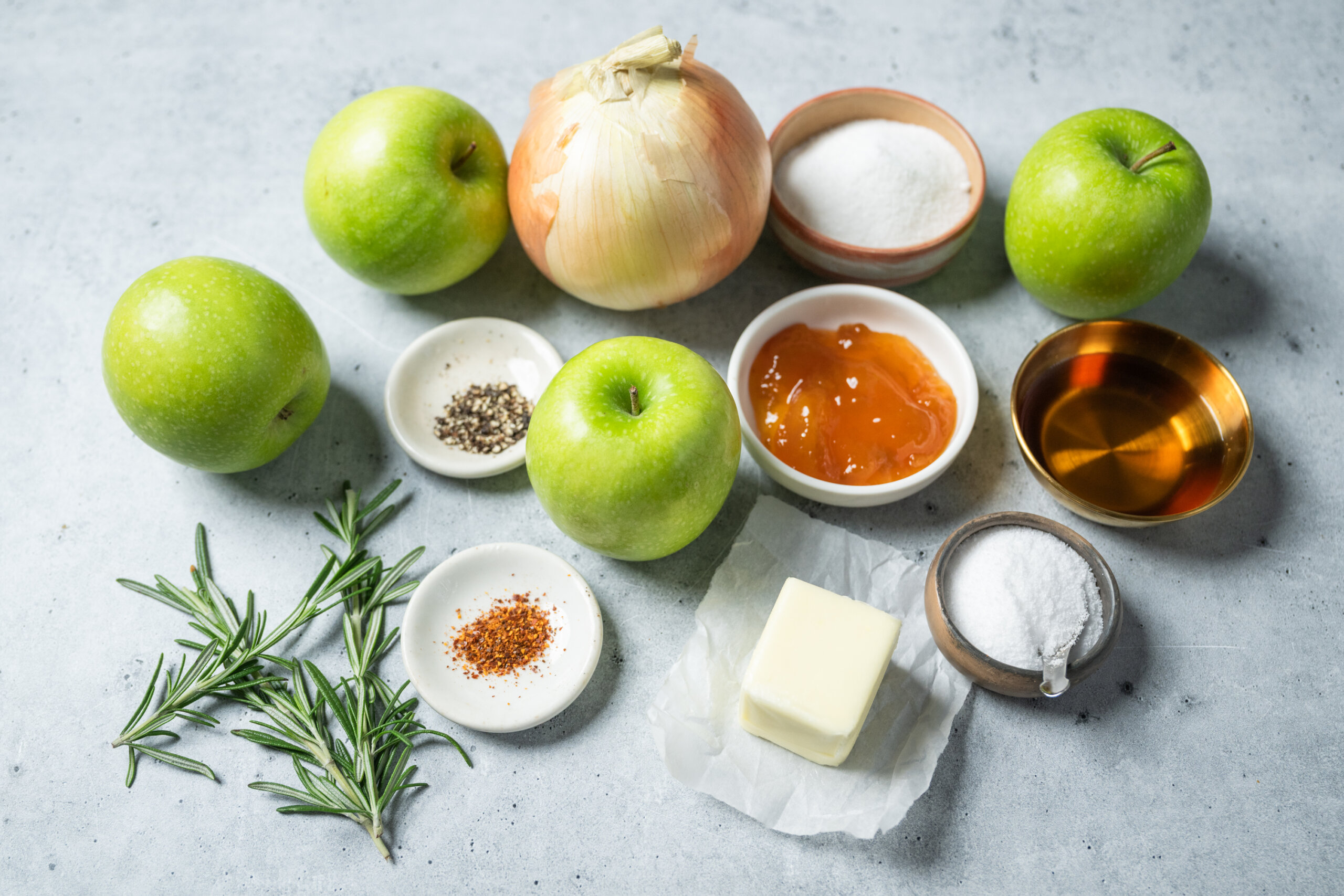 Green apples, a yellow onion, butter, rosemary, salt, pepper, chili flakes, granulated sugar, apricot jam, and a small bowl of liquid arranged on a gray surface.