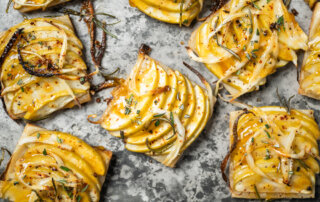 Rectangular slices of puff pastry topped with thinly sliced Granny Smith apples, onions, herbs, and seasoning on a textured gray surface.