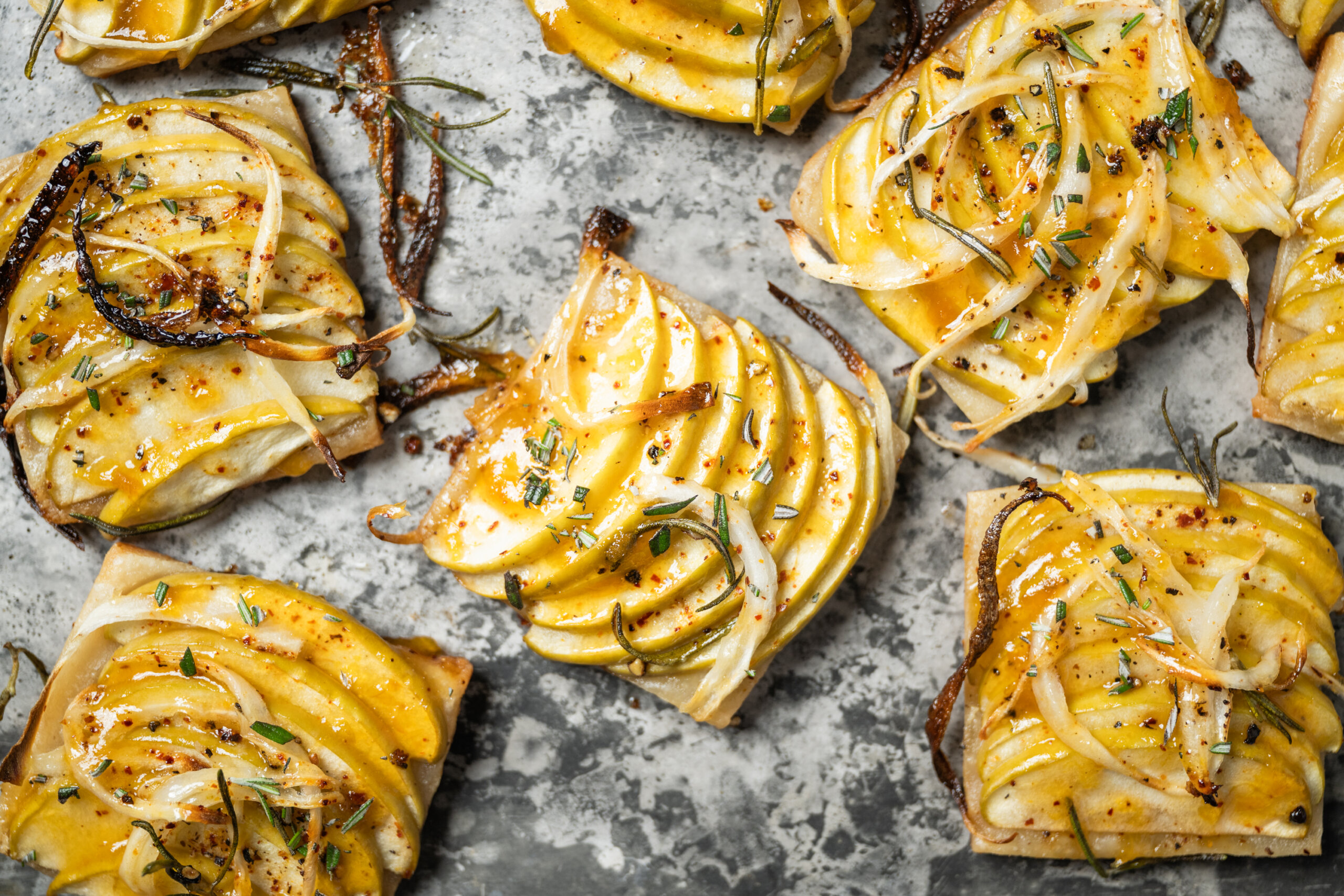 Rectangular slices of puff pastry topped with thinly sliced Granny Smith apples, onions, herbs, and seasoning on a textured gray surface.