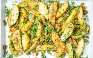 Roasted potato wedges on a white tray, garnished with chopped parsley and sliced green olives.