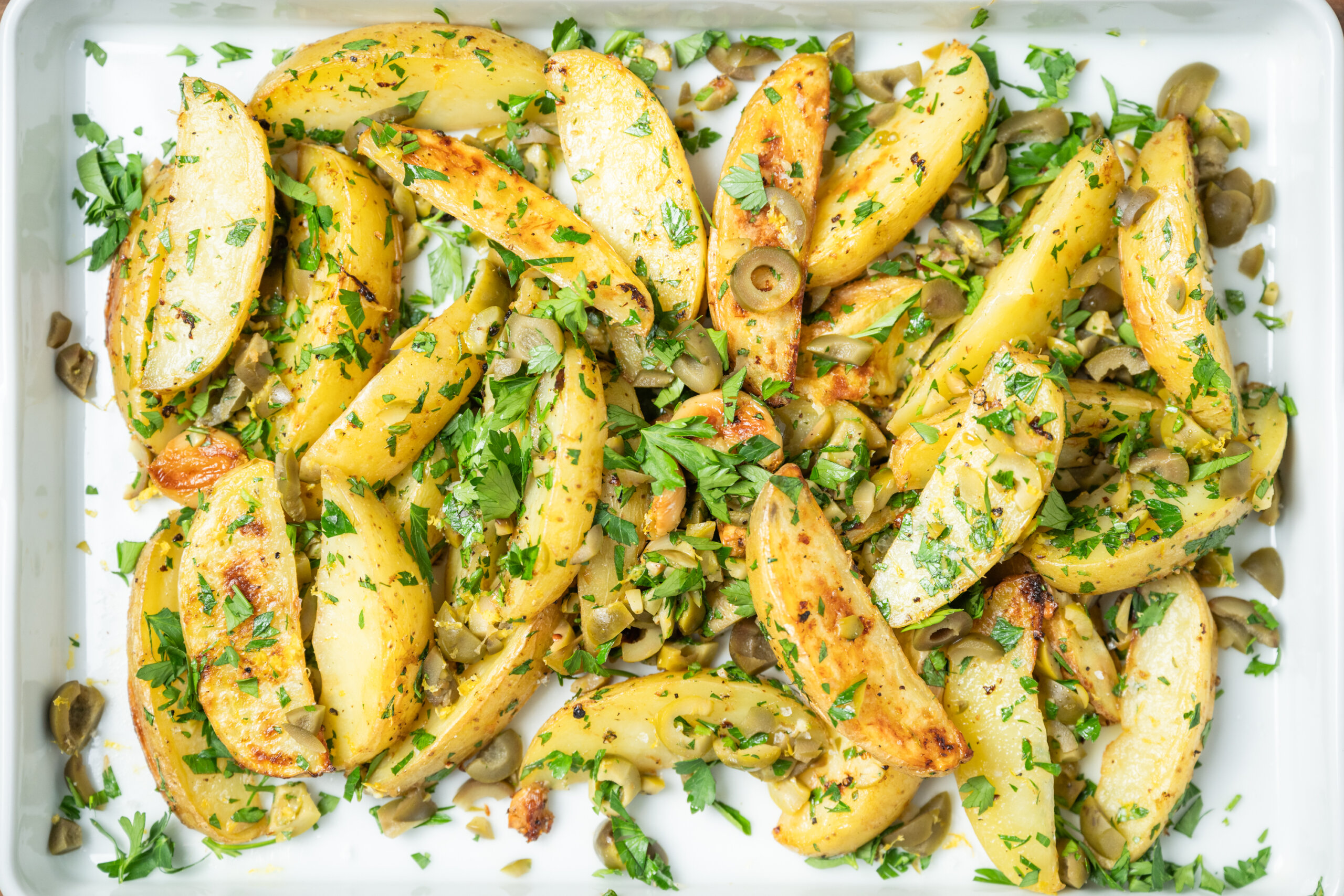 Roasted potato wedges on a white tray, garnished with chopped parsley and sliced green olives.