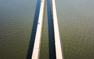 Aerial view of a long, two-lane bridge with parallel spans crossing a large body of water, with minimal traffic and clear shadows on the water's surface.