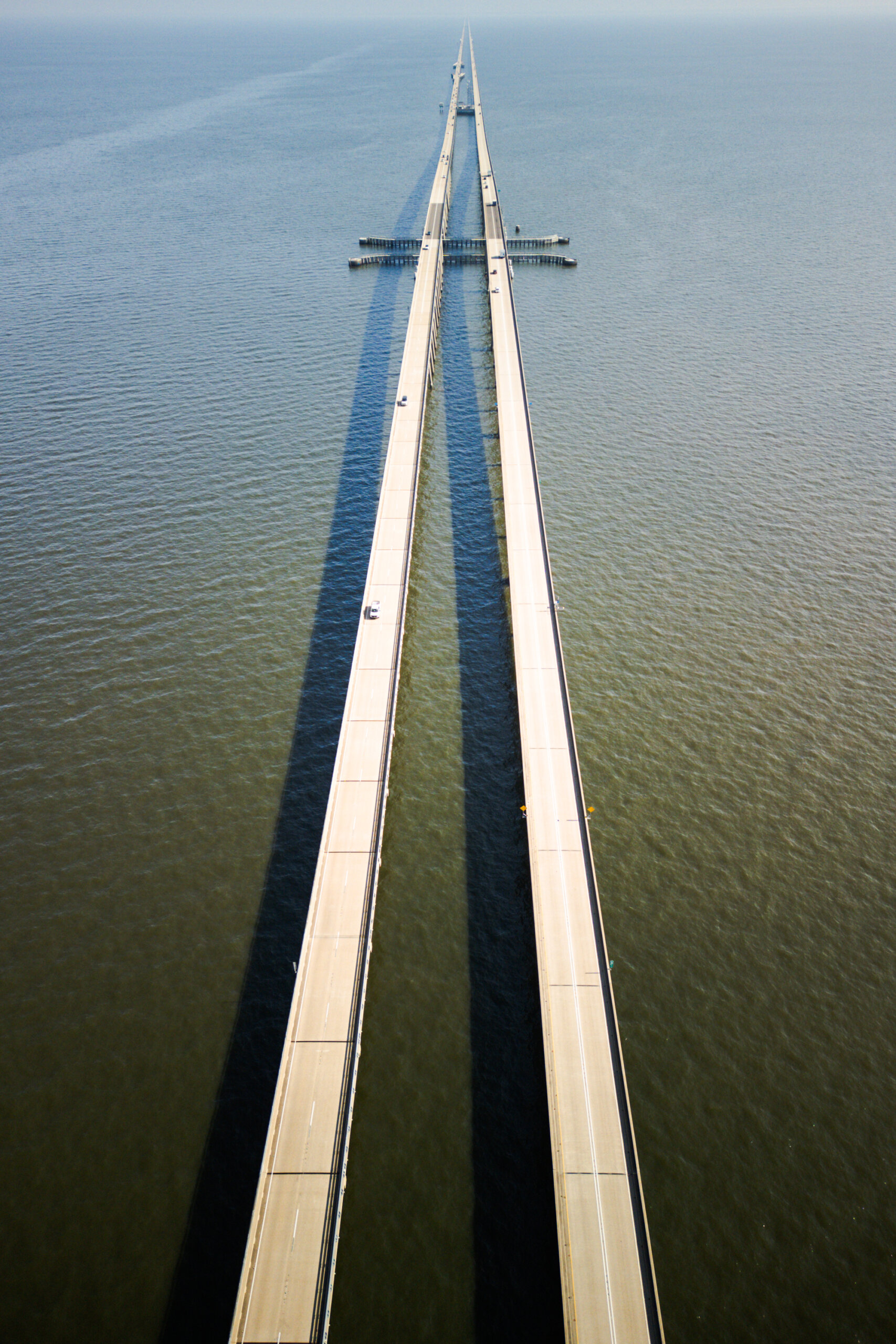 Aerial view of a long, two-lane bridge with parallel spans crossing a large body of water, with minimal traffic and clear shadows on the water's surface.