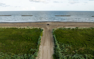 A sandy road leads from green vegetation to a beach with gentle waves and rock barriers in the water; a few people and a vehicle are on the sand near the shoreline.