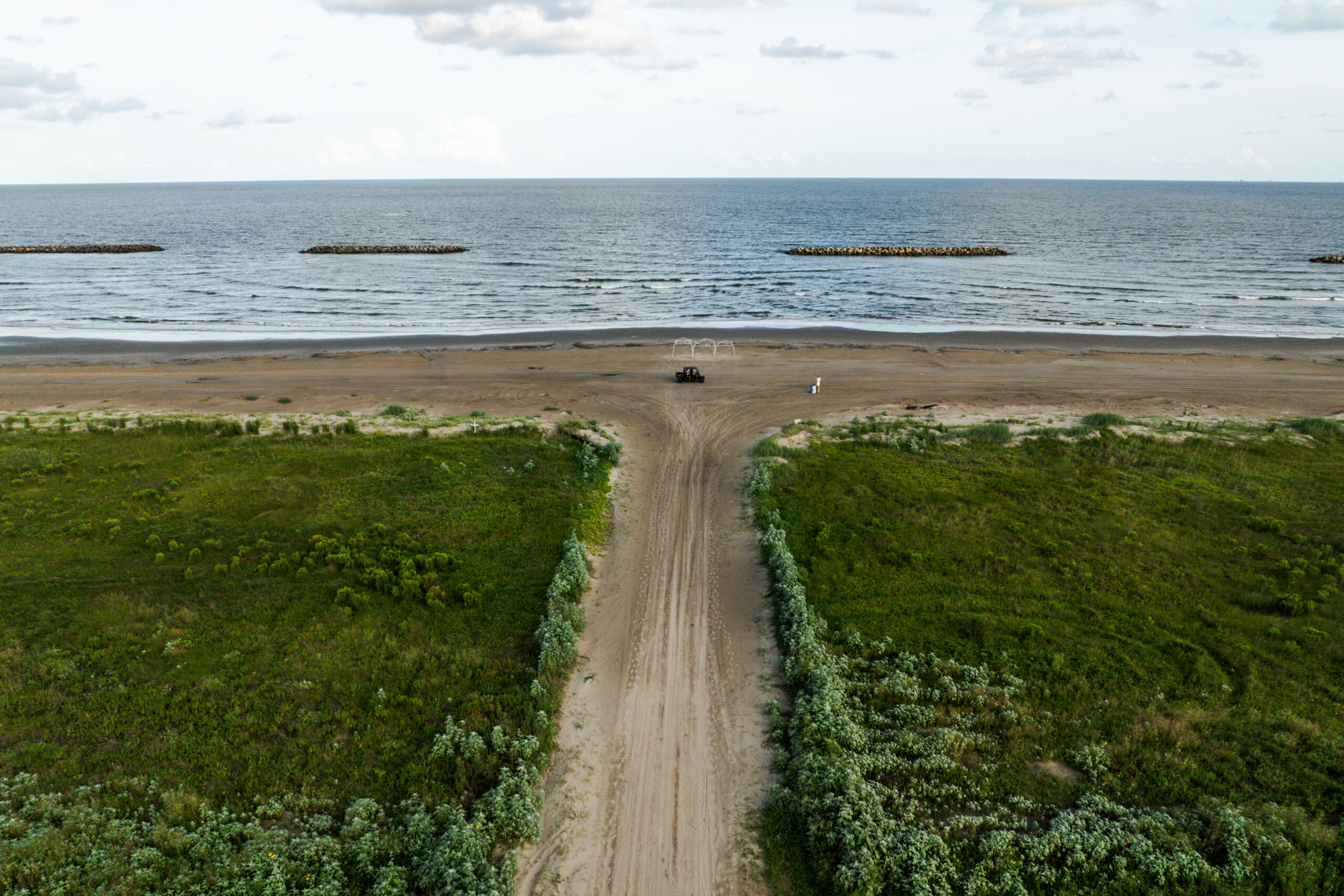 A sandy road leads from green vegetation to a beach with gentle waves and rock barriers in the water; a few people and a vehicle are on the sand near the shoreline. A sandy road leads from green vegetation to a beach with gentle waves and rock barriers in the water; a few people and a vehicle are on the sand near the shoreline.