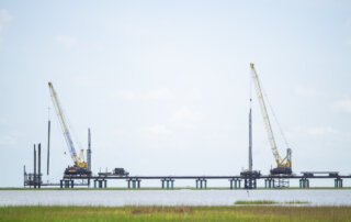 Two large cranes are positioned on a partially constructed bridge over water, with grassy land in the foreground and a clear sky above.