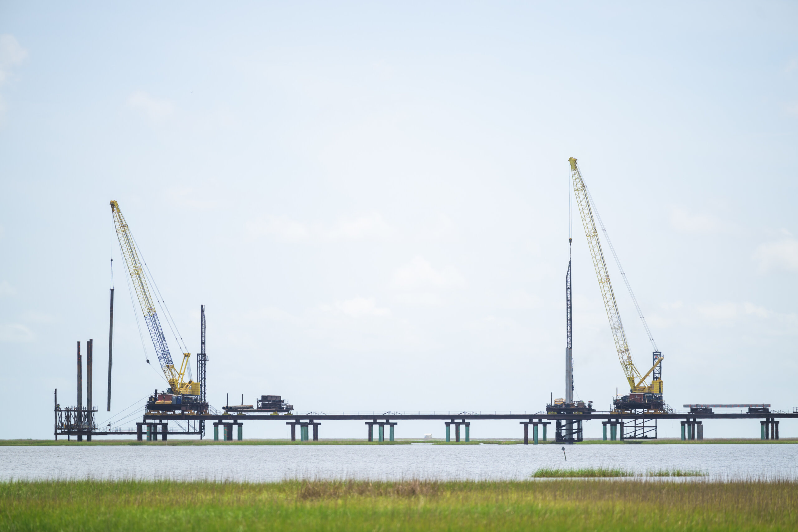 Two large cranes are positioned on a partially constructed bridge over water, with grassy land in the foreground and a clear sky above. Two large cranes are positioned on a partially constructed bridge over water, with grassy land in the foreground and a clear sky above.